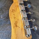 Detail zadní strany hlavy kytary z přírodního dřeva. Na dřevě je vyryt nápis „Key to the Rock“. Jsou vidět kovové ladicí mechanismy v chromovaném pro…
