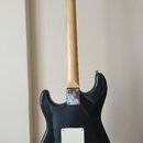 Zadní strana černé elektrické kytary s jasným dřevěným hřednem. Na zadní straně hlavy je nápis Squier by Fender a nečitelný štítek. Na spojení hřdnu…