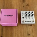 Růžové papírové krabička s nápisy GOLDEN MASTER PEDAL a ENDORPHINES. Vedle ní je kytarový efekt s bílým povrchem, který nese název GOLDEN MASTER PEDA…
