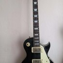 Černá elektrická kytara s bílým pickguardem a zlatými prvky. Na hlavě hředu je nápis Vintage a symbol lilie. Nástroj má dva humbuckery, čtyři otočné…