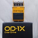 Žlutý kytarový efekt s nápisy OverDrive a OD-1X oraz logem BOSS na černém spínacím pedálu. V horní části jsou čtyři ovládací knoflíky a nápisy OUTPUT…