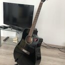 Černá akustická kytara s výřezem v korpusu, umístěná na černém stojanu. Na hlavě hředu je viditelné logo Fender. Kytara má tmavý hmatník a červený tr…
