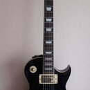 Černá elektrická kytara s tvarem typu Les Paul. Na hlavě je bílý nápis Vintage a pod ním symbol lilie. Kytara má dva humbuckery, čtyři ovládací knofl…