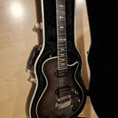 Tmavě hnědá elektrická kytara s dvojstupňovými humbuckery a bílým lemováním těla. Nástroj je umístěn v otevřeném černém tvrdém kufru s bílým okrajem.…