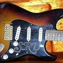 Elektrická kytara s tělem v provedení sunburst a černým pickguardem. Na pickguardu je bílý nápis v ozdobném písmu SRV. Kytara má tři snímače s bílými…