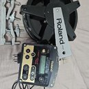 Na fotografii je černý elektronický pad s kovovou lištou, na které je nápis Roland PD-85. Vedle něj je černý trigger modul s gold detaily a nápisy Ro…