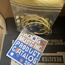 V kartonové krabici se nachází průhledný plastový sáček s žlutými patch kabely. Před ním leží modrý papírový katalog s nápisem MOOG MUSIC INC. PRODUC…