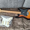 Elektrická kytara s tělem v provedení sunburst a hnědým hřebenatým pickguardem. Na hlavě je viditelné logo Washburn. Vedle kytary leží v plastovém ob…