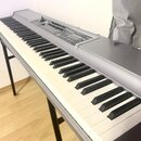 Digitální klavír v šedé barvě s černými a bílými klávesami, umístěný na černém kovovém stojanu. Horní panel obsahuje ovládací prvky, knoflíky a malý…