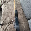Fotografie zachycuje elektrickou kytaru z profilu. Nástroj má černý lakovaný korpus a světle hnědý dřevěný krk. Na kytaru je instalován tremolo systé…
