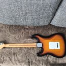 Zadní strana elektrické kytary s tónem sunburst (přechod od černé k žluté). Kytara má světle hnědý krk a bílou plastovou krytku elektroniky na zadní…