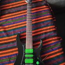 Černá elektrická kytara s třešňovým hnedým hmatníkem a třemi zelenými humbuckery. Na hlavě nástroje je viditelné logo, které není čitelné. Kytara má…