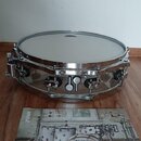 Stříbrný kovový buben s bílým blátcem. Na doprovodném letáku je vidět logo SONOR a nápisy SQ2 oraz DRUM SYSTEM. Nástroj vykazuje čistý, lesklý povrch…