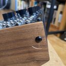 Detailní záběr na hranu hudebního zařízení s dřevěným povrchem v hnědé barvě. Na horní hraně jsou vidět černé ovládací knoflíky a řada kovových konek…