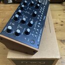 Černý analogový syntetizátor Moog MOTHER-32 v dřevěném šasi, umístěný na kartonové krabici s nápisem moog MOTHER-32. Přední panel obsahuje numerous o…