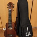 Hnědý dřevěný ukulele s černým pouzdrem. Na hlavě nástroje i na přední straně pouzdra je bílé logo znázornující korunu nad písmenem M. Uvnitř otvoru…