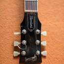 Fotografie zachycuje hlavu a část hmatníku elektrické kytary v černé barvě. Na hlavě jsou bílé nápisy „Standard“, „Les Paul“ a logo „Gibson“. Jsou vi…