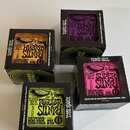 Pět krabiček kytarových strun značky ERNIE BALL. Každá krabička má jinou barvu a označení modelu: HYBRID SLINKY (oranžová), POWER SLINKY (fialová), S…