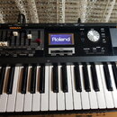 Černý digitální klavír s bílými a černými klávesami. Na ovládacím panelu je modrý displej s bílým nápisem Roland a oranžové nápisy ORGAN a PIANO. Pan…