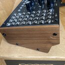 Černý analogový syntetizátor s ovládacími knoflíky a řadou patchových konektorů, umístěný v dřevěném šasi. Na spodní části kartonové krabice je bílý…