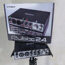 Fotografie zobrazuje USB audio rozhraní v černé barvě umístěné na stojanu, vedle něj je originální krabice. Na zařízení i na krabici je vidět logo Ro…