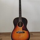 Akustická kytara s korpusem v barvě sunburst a tmavým hmatníkem. Nástroj je umístěn na černém stojanu před světlou stěnou. Na hlavě kytary jsou vidět…