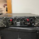 Zadní strana černého rackového zesilovače s dvěma velkými ventilátory po stranách. Na panelu jsou nápisy CHANNEL B OUT 4Ω/600W, BRIDGE OUT 8Ω/1200W a…