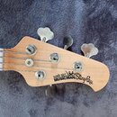 Fotografie zachycuje hlavu basové kytary z přírodního světlého dřeva s kovovými ladičkami. Na dřevě je černý nápis ERNIE BALL MUSICMAN StingRay. Povr…