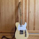 Světle žlutá elektrická kytara typu Telecaster s bílým pickguardem a javorovým hmatníkem. Na hlavě krku je viditelné logo Fender. Kytara má kovový mo…