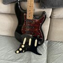 Černá elektrická kytara s hnědým javorovým hmatníkem a červeně vzorovaným pickguardem. Vedle kytary leží samostatný černý pickguard s třemi bílými sn…