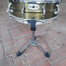 Zlatavý snare drum s kovovými obručemi umístěný na chromovaném stojanu. Na korpusu je oválný štítek s nápisy Pearl a SENSITONE. Na horním obruči je č…