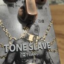 Detail efektového pedálu s kovovým povrchem a třemi hnědými ovládacími knoflíky. Na spodní části je nápis TONE SLAVE a logo MH pedals. Vedle knoflíků…