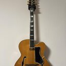 Polodublová elektrická kytara v žlutohnaté barvě s černým hmatníkem a černým plechtrem. Na hlavě je bílý nápis Epiphone a ozdobné vložky. Nástroj má…