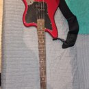 Červená elektrická basová kytara s černým pickguardem a černým popruhem. Na hlavě nástroje je bílý nápis Squier by Fender JAGUAR BASS. Povrch nástroj…