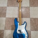 Modrá elektrická basová kytara s bílým pickguardem a hnedým hmatníkem. Na hlavě nástroje je viditelné logo Fender. Povrch laku je glossy, na těle jso…