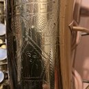 Detail zlatavého kovového povrchu hudebního nástroje s viditelným opotřebením a drobnými škrábancemi. Na povrchu je vyryto logo s podobou ženské tvář…