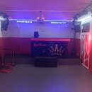 Interiér místnosti s červenými stěnami a šedou podlahou. V centru je pult s nápisem RED VELVET a grafikou koruny, pod ním je umístěn černý subwoofer.…