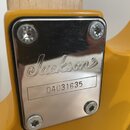Detail kovového krytu na žlutém těle kytary. Na krytu je vyryto logo Jackson® a pod ním v rámečku sériové číslo 04031635. Kovový povrch je lesklý a v…