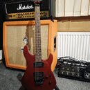 Tmavě červená elektrická kytara s hnědým hmatníkem a černými snímači. Na hlavě je bílý nápis Ibanez. V pozadí je kytarový zesilovač Marshall s nápise…