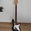 Černá basová kytara s bílým pickguardem a hnědým hmatníkem. Na hlavě nástroje je viditelné logo Fender a nápis PRECISION BASS. Nástroj stojí na černé…