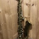 Mosazný saxofon s hnědým mouthpiecem. Nástroj vykazuje známky opotřebení povrchu a patinu v několika místech. Na těle nástroje nejsou viditelné žádné…
