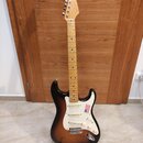 Elektrická kytara s tvarem Stratocaster, v barevné variantě sunburst a s bílým pickguardem. Na hmatníku a hlavě je vidět světlý dřevěný materiál, na…