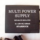 Černý hranatý zdroj napájení s textem MULTI POWER SUPPLY, SEPARATE ISOLATED a 6 x 10V DC/100mA. Dole je uveden nápis STARBUCK. Povrch zařízení má jem…