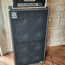 Fotografie zobrazuje basový reproduktor Ampeg s černou síťovkou a logem Ampeg v levém horním rohu. Na reproduktoru stojí rack s vybavením, včetně zař…