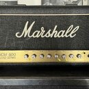 Kytarový zesilovač značky Marshall, model JCM 800 LEAD SERIES. Má černý kryt s bílým logem Marshall a zlatý ovládací panel s řadou otočných knoflíků.…
