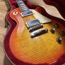 Elektrická kytara s trupem v barvě sunburst a viditnou strukturou dřeva, umístěná v otevřeném kufru s červeným vnitřkem. Nástroj má dva humbuckery, k…