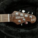 Fotografie zachycuje hlavu elektrické kytary z hnědého dřeva s chromovanými ladičkami. Na povrchu je bílý nápis ERNIE BALL MUSIC MAN® a logo LIII™. P…