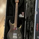 Elektrická kytara s tmavě šedým metalizovaným povrchem v černém tvrdém kufru. Na hlavě hmatníku je nápis MUSIC MAN a na boku kufru červený nápis MUSI…