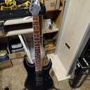 Černá elektrická kytara s tmavým hnedým hmatníkem, umístěná na stojanu. Na hlavě nástroje je bílý nápis Ibanez. Kytara má dva humbuckery a černý povr…