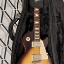 Elektrická kytara s tmelem v barvě sunburst uložená v černém plišovém pouzdře. Na hlavě hmatníku a na černé oporce je bílý nápis Gibson. Kytara má dv…